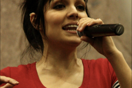 Headshot of a woman with brown hair wearing a red shirt and holding a microphone