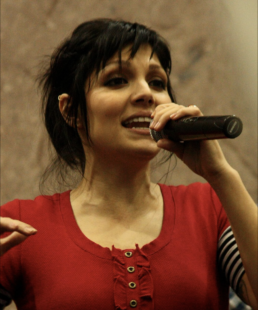 Headshot of a woman with brown hair wearing a red shirt and holding a microphone